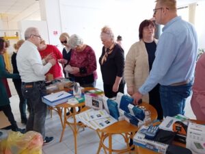 Na stoloch s sú husto poukladané rôznorodé kompemzačné pomôcky. Pred stolmi v rade stoja účastníci prednášky, ktorí spoznávajú jednotlivé pomôcky. Pán Červenka vysvetľuje.