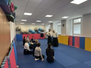 V telocvični, v tvare polkruhu na žinenkách sedia účastníci športového krúžku. Pred nimi stojí dvojica inštruktorov oblečených v Aikido oblečení - široké nohavice a voľná košeľa. Názorne predvádzajú a popisujú cvičenia.